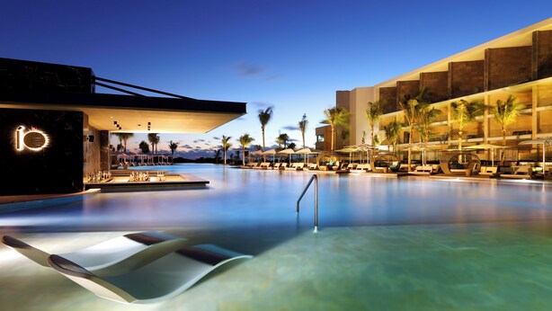 A modern hotel with a large infinity pool, palm trees, and a well-lit facade.