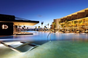 Een modern hotel met een groot overloopzwembad, palmbomen en een goed verlichte gevel.