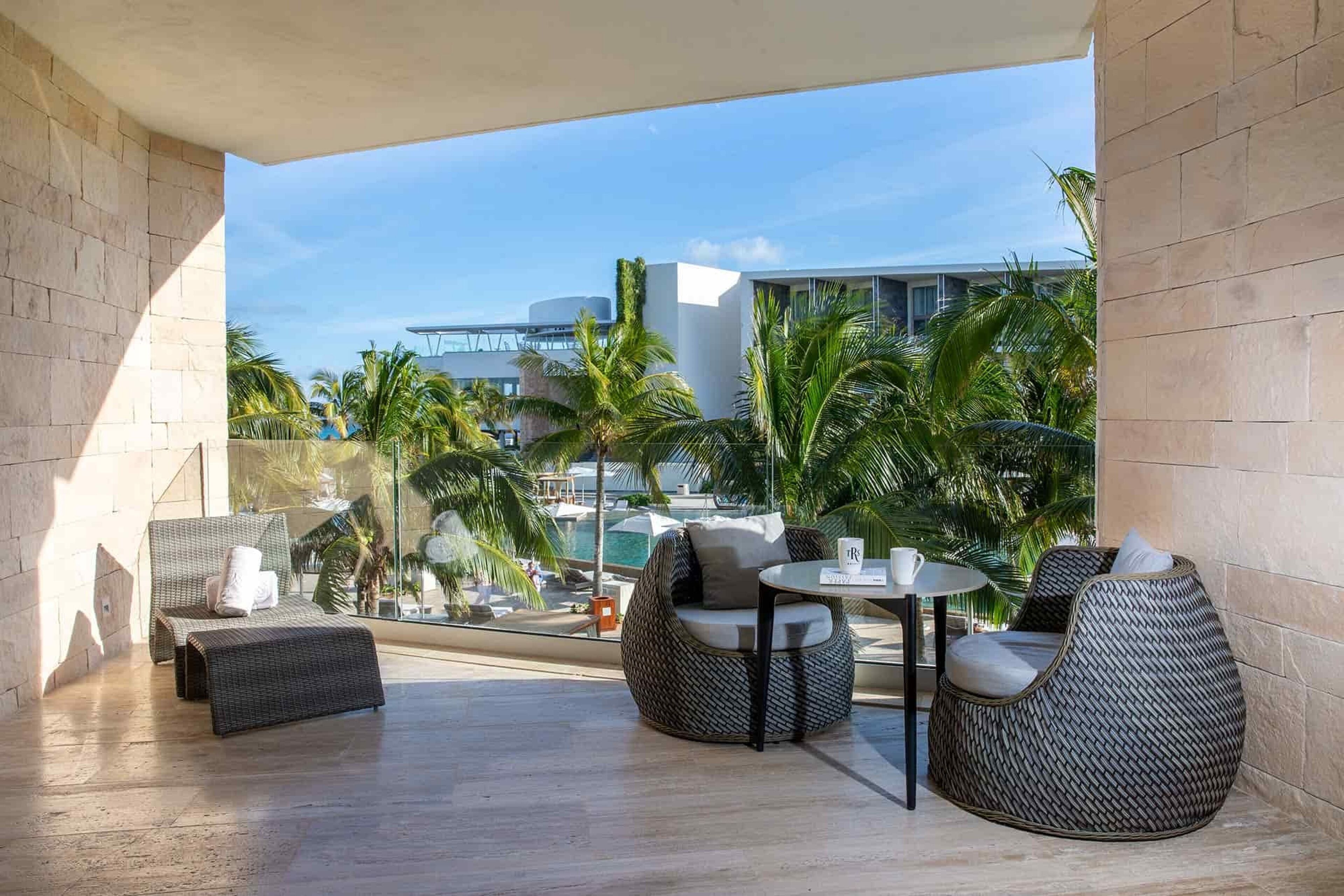 A modern outdoor lounge area with wicker furniture and a glass balustrade overlooking a pool and palm trees.