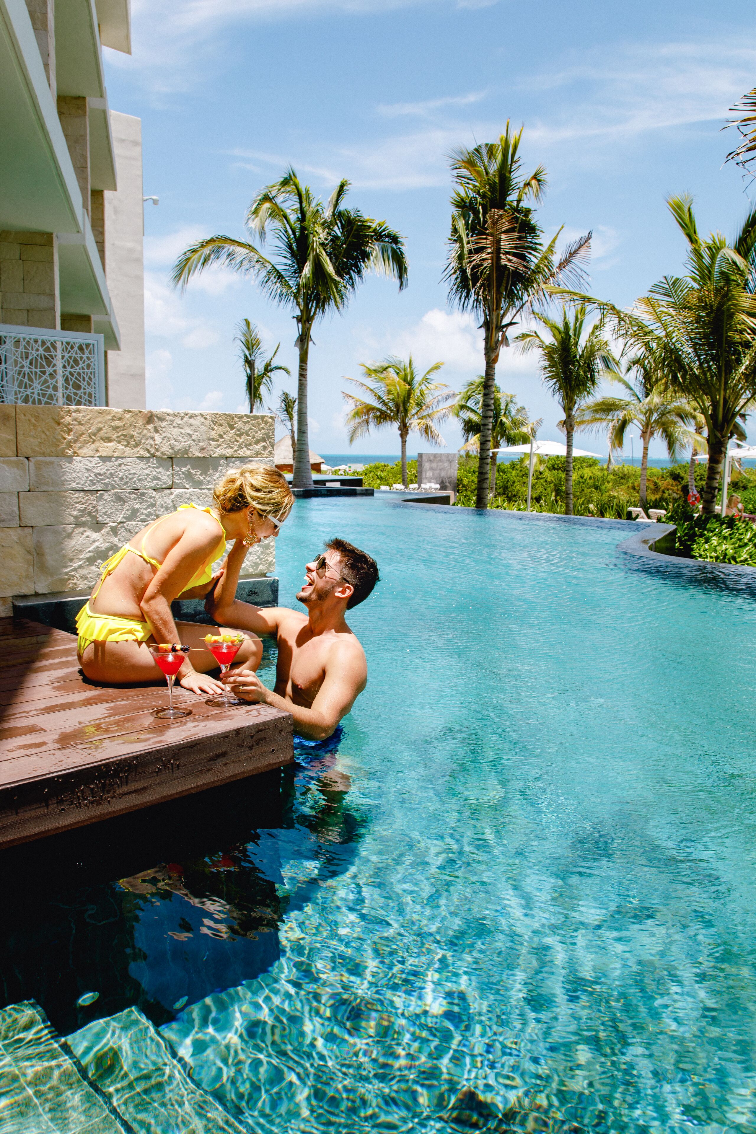 A couple by a pool with clear water, surrounded by palm trees and a stone wall.