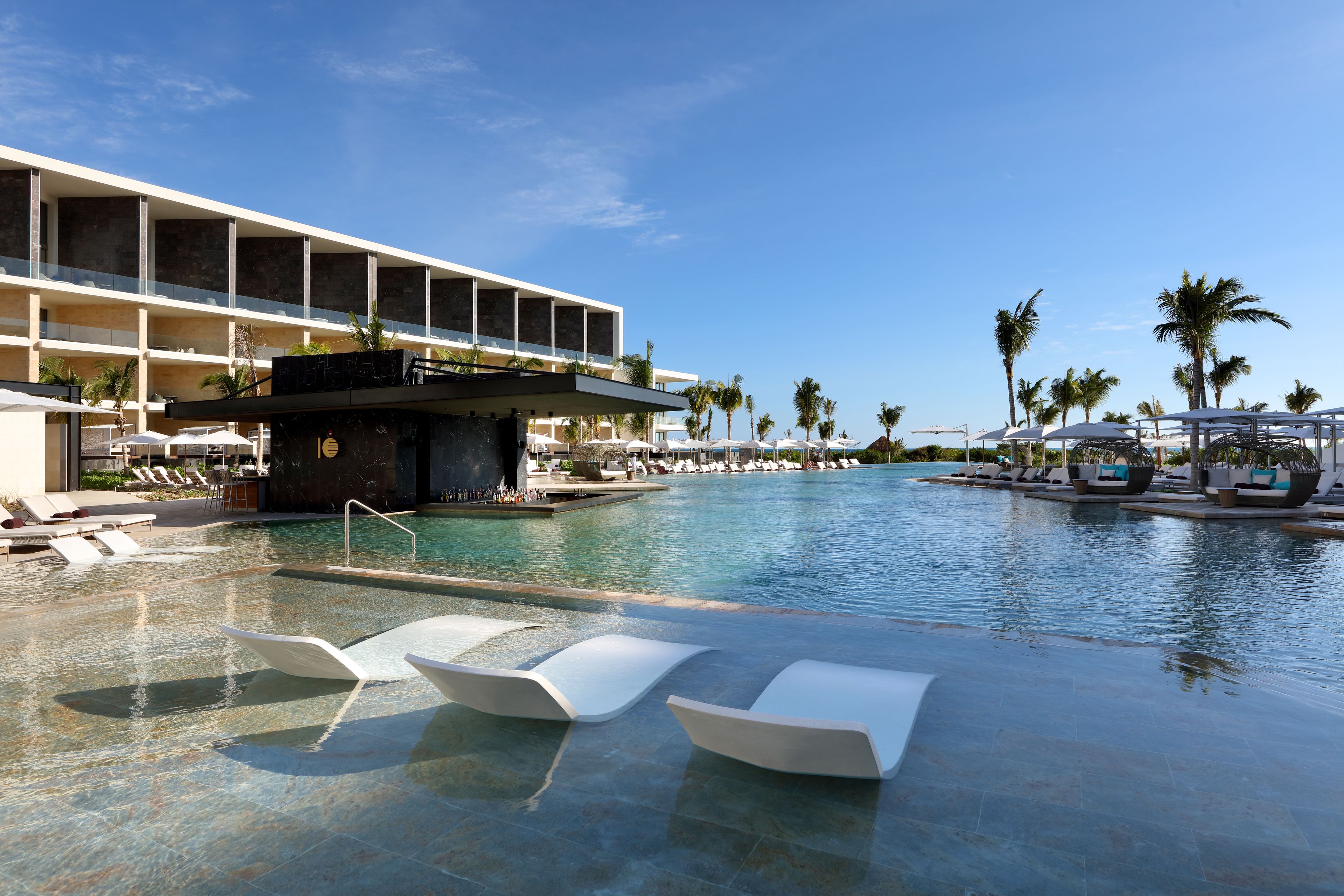 A modern hotel with a large infinity pool, sun loungers, and palm trees.