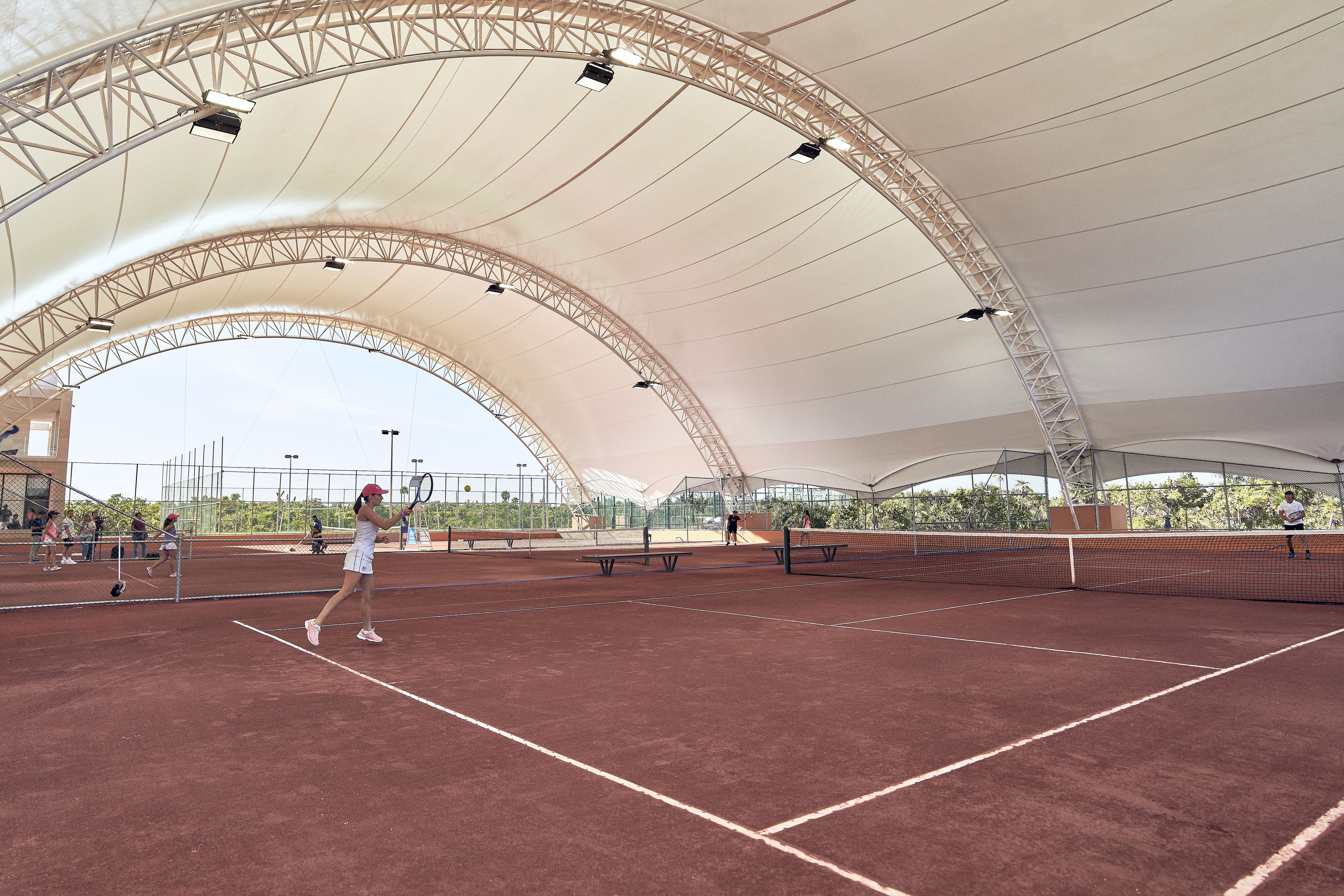 An indoor tennis court with a high, arched roof and multiple lighting fixtures.