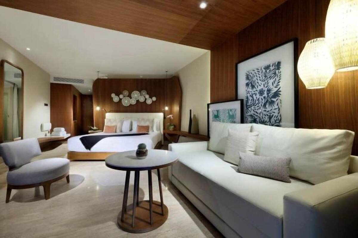 A modern hotel room with a bed, a sofa, a round table, and a wall-mounted light fixture.
