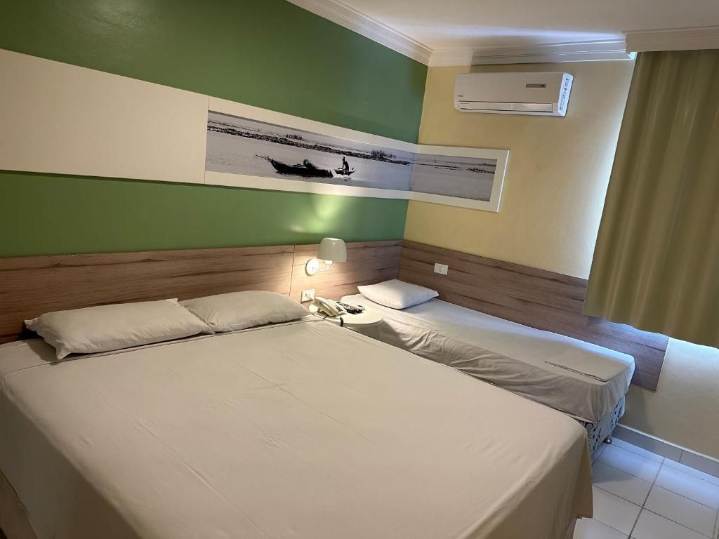 A hotel room with a large bed, a smaller bed, a nightstand with a lamp, and a wall-mounted air conditioner.