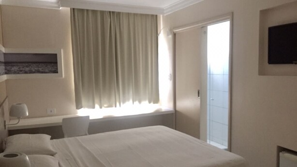 A hotel room with a bed, a desk, a chair, a TV, and a window with curtains. - Hotel Porto Grande (Maceió)