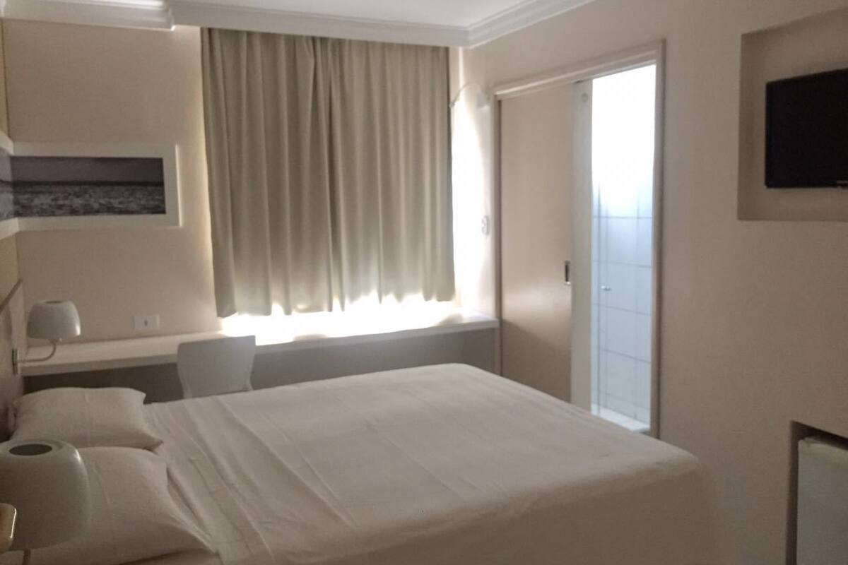 Habitación de hotel con cama, escritorio, silla, televisión y ventana con cortinas.