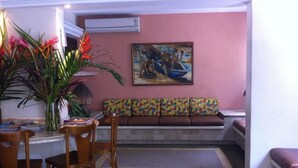 A room with a tiled floor, a bench with colorful cushions, a painting on the wall, and a plant with red flowers. - Hotel Porto Grande (Maceió)