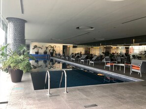 A modern indoor pool area with lounge chairs and tables, surrounded by potted plants and a large pillar. - Plaza Suites México City 2404 (Mexico City)