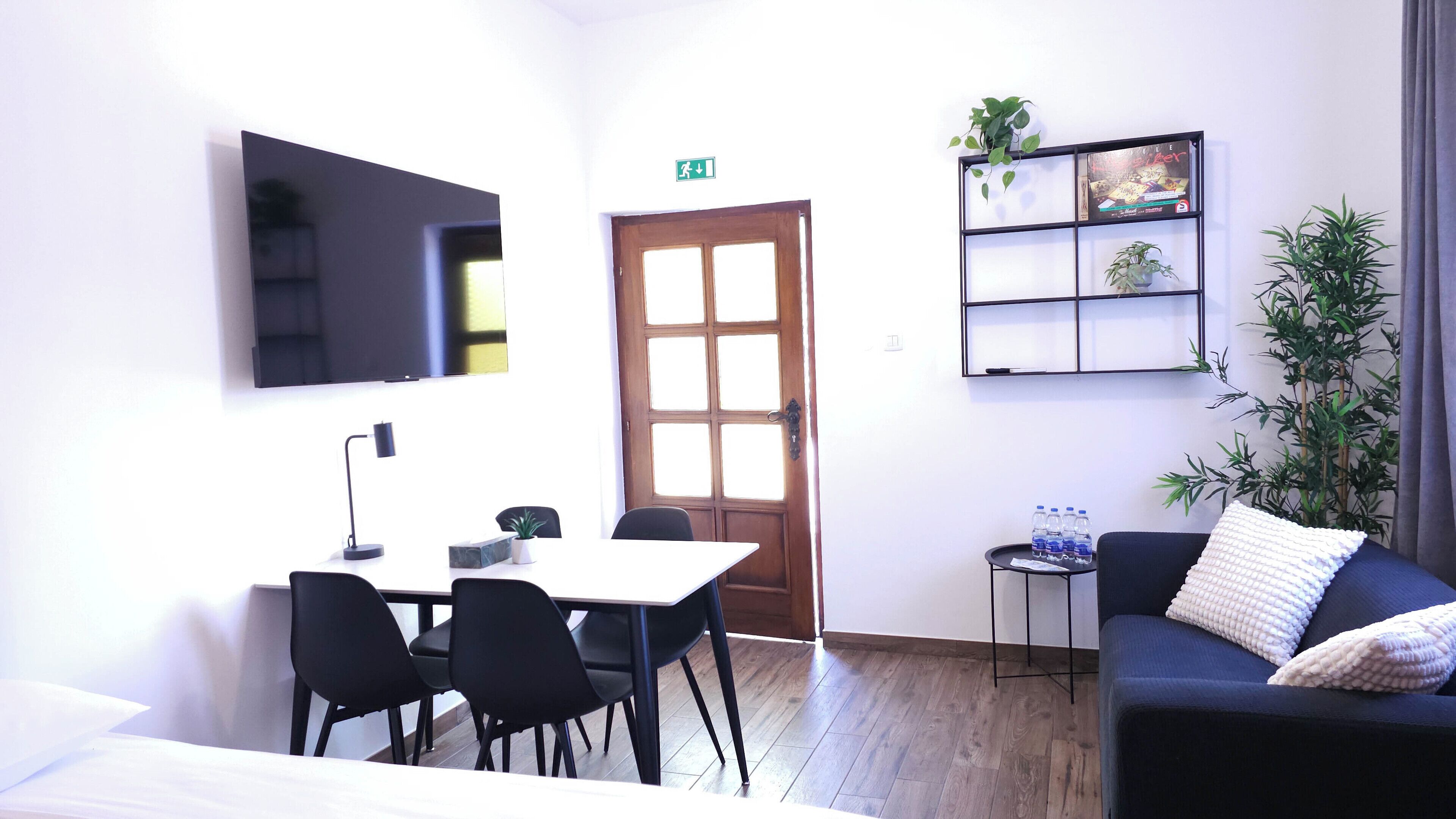Habitación moderna con puerta de madera, televisor montado en la pared, una mesita con sillas, un sofá con cojines y una estantería con plantas y una botella de agua.