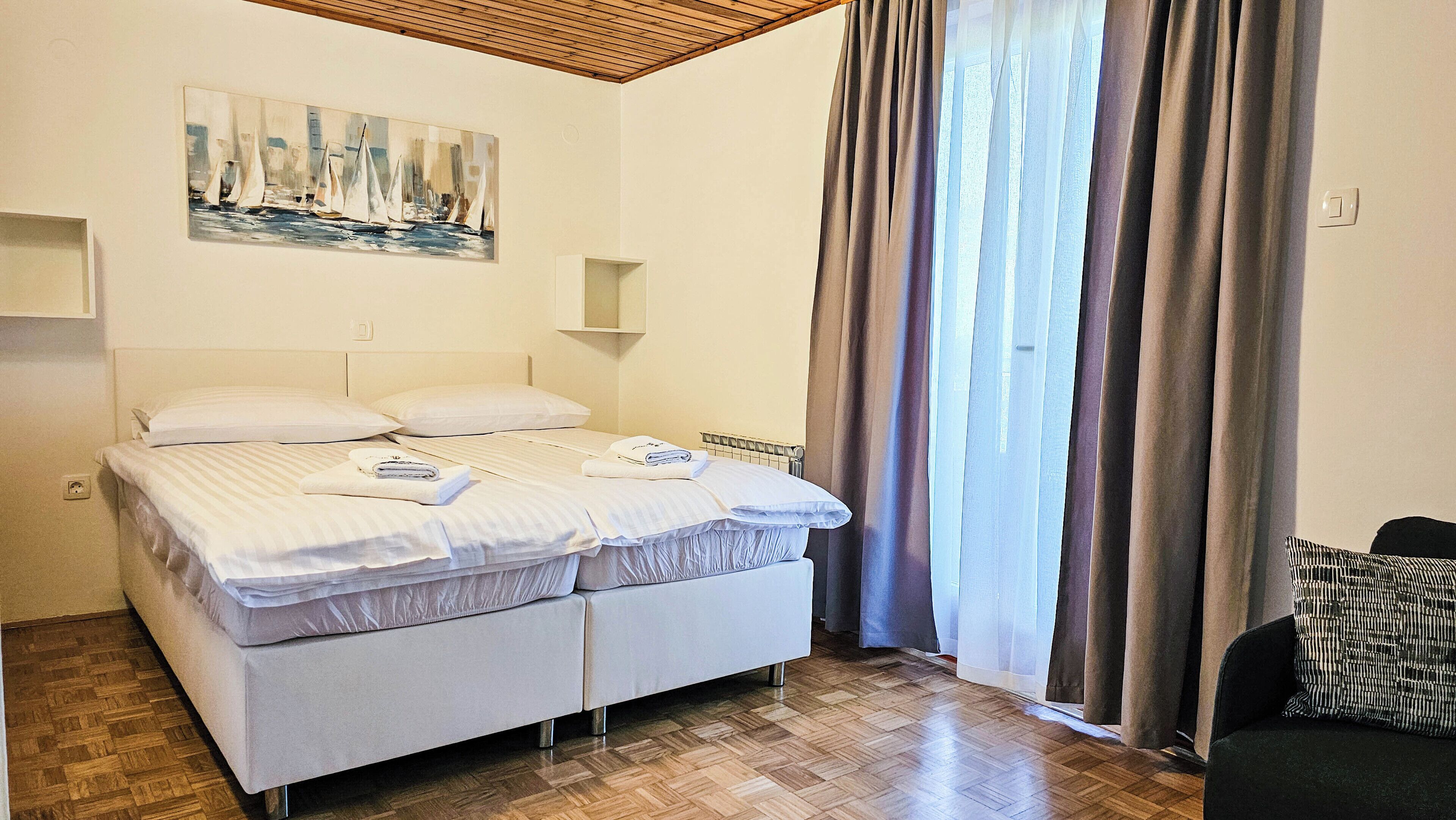 Ein Doppelbett mit weißer Bettwäsche, ein Segelbootsgemälde an der Wand, ein Fenster mit Vorhängen und ein schwarzer Sessel.