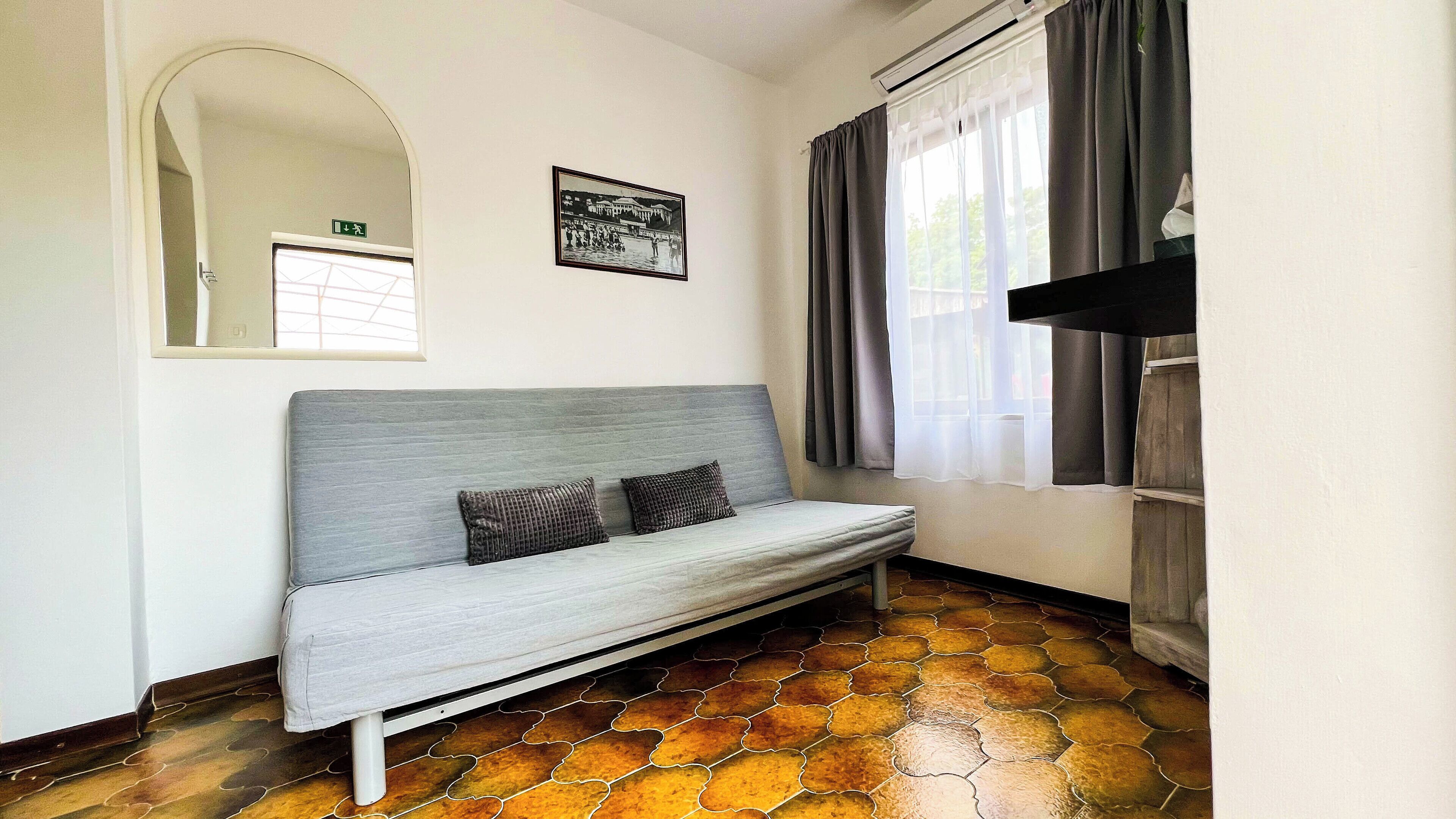 Una habitación con un sofá gris, un espejo, un cuadro enmarcado y una ventana con cortinas.
