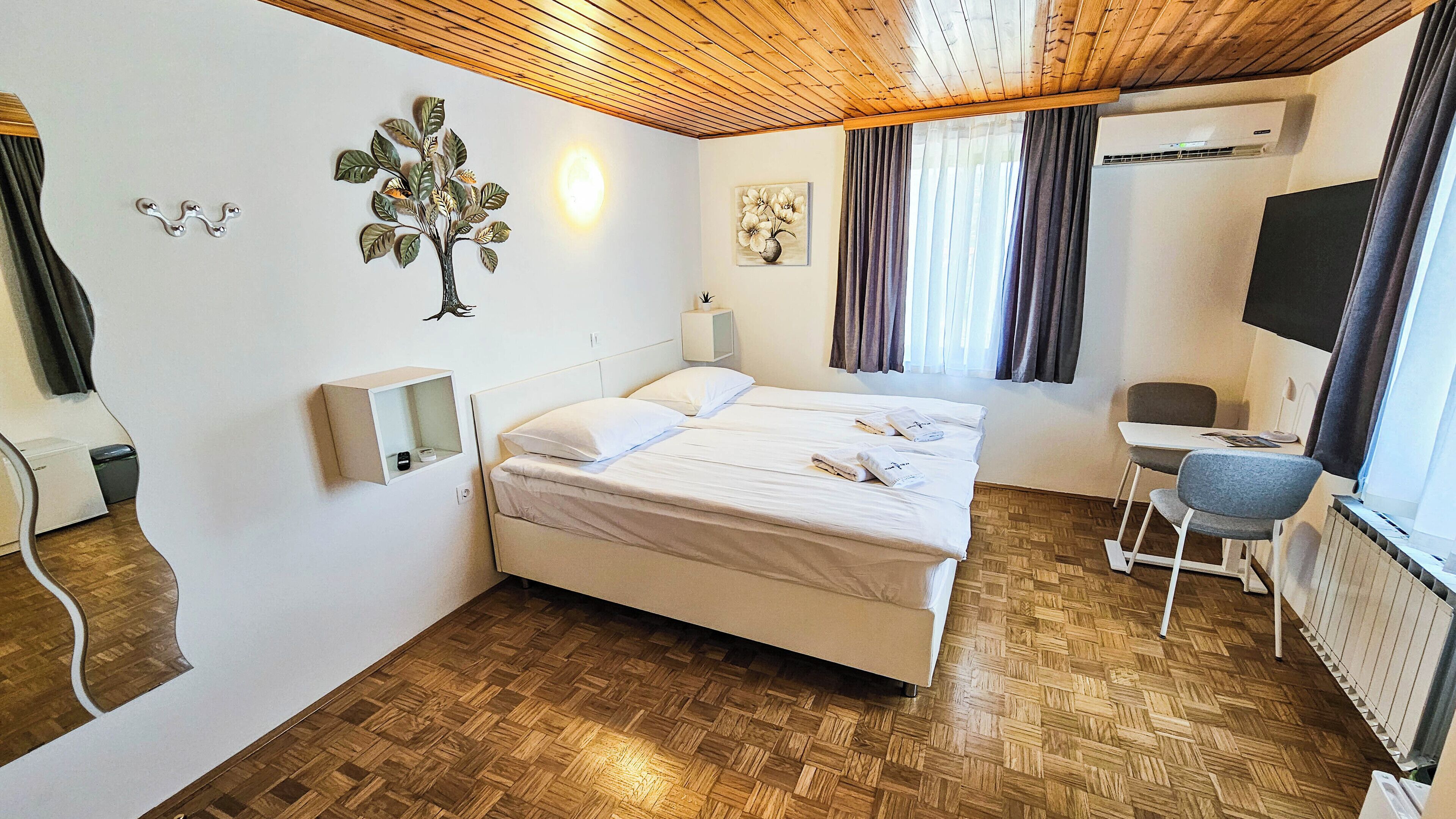 Ein Hotelzimmer mit Holzboden, einem Bett mit weißer Bettwäsche, einem Schreibtisch mit Stuhl, einer Wandklimaanlage und einem Fenster mit Vorhängen.