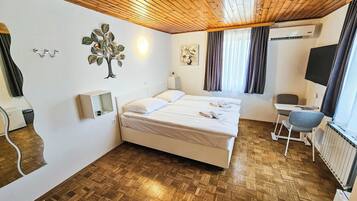 Ein Hotelzimmer mit Holzboden, einem Bett mit weißer Bettwäsche, einem Schreibtisch mit Stuhl, einer Wandklimaanlage und einem Fenster mit Vorhängen.