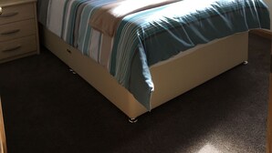 Een slaapkamer met een bed, een nachtkastje, een lamp, een raam met gordijnen, een schilderij aan de muur en een bloevas op de vensterbank.