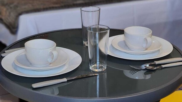 A neatly made bed with white linens and a gray throw blanket, a bedside table with a lamp, and a small round table with a glass of water, two cups, and cutlery. - Bonito Hi Hostel e Pousada (Bonito)