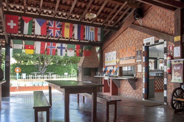 An outdoor dining area with tables and benches, a brick wall, and a sign for 'Cozinha Kitchen'. Flags are hanging above. - Bonito Hi Hostel e Pousada (Bonito)
