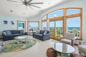 A spacious living room with a large window offering a view of the ocean, a ceiling fan, and a blue sofa with a glass coffee table. - Heaven By The Sea: Oceanfront, Pool, Spa, Elevator (Nags Head)