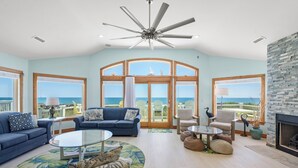 Un salon spacieux doté d’un grand ventilateur de plafond, d’un canapé bleu, d’une table basse en verre et offrant une vue sur l’océan grâce à de grandes fenêtres.