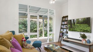 Ein modernes Wohnzimmer mit einem Glastisch, einer Couch mit bunten Kissen, einem Bücherregal und einem großen Fernseher, der eine Waldszene zeigt.