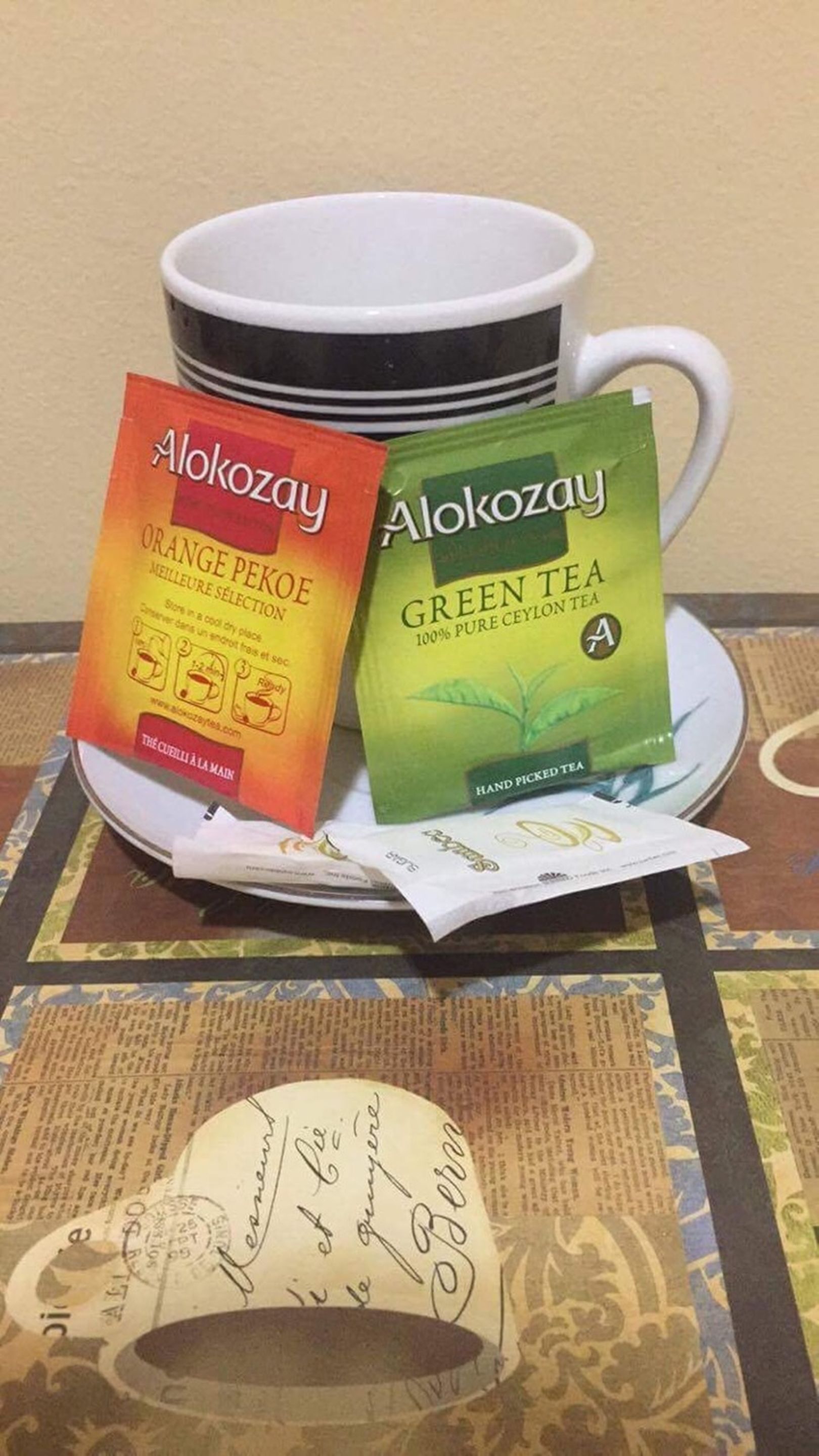 Two Alokozay tea packets, one orange pekoe and one green tea, placed on a saucer next to a white mug.