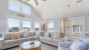 Ein geräumiges Wohnzimmer mit einer großen Eckcouch, einem Glastisch und Blick auf benachbarte Häuser durch große Fenster.