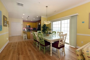 A dining area with a table and chairs, a kitchen in the background, and a large window with curtains.