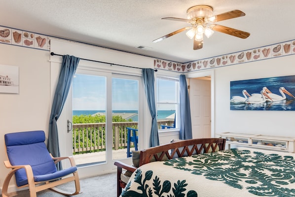 A bedroom with a bed, a rocking chair, a ceiling fan, and a balcony with a view of the ocean. - Crab Cottage: Oceanfront, Private Pool, Hot Tub (Nags Head)