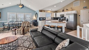 Ein großzügiges Wohnzimmer mit einer schwarzen Ledersofa, einem Holztisch und einem gemusterten Teppich. Es gibt einen Essbereich mit Stühlen und einem Tisch, sowie eine Küche mit modernen Geräten und weißen Schränken.