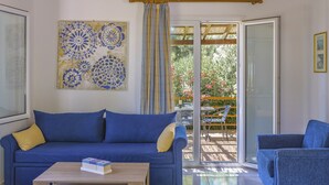 Ein Zimmer mit einem blauen Sofa, einem Holztisch, einem blauen Sessel, einem Wandbehang mit Muster und Blick auf eine Terrasse mit Tisch und Stühlen.