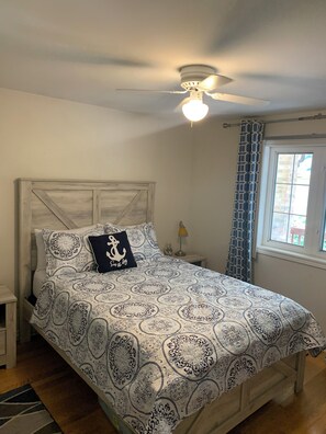 A bedroom with a bed, a ceiling fan, a window with curtains, and a nightstand with a lamp.