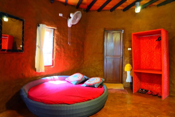 Una habitación con cama redonda, un ventilador, un espejo, una ventana con cortinas, una puerta de madera y un estante rojo con zapatos.