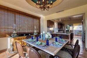 A dining area with a table set for four, surrounded by wicker chairs, a chandelier, and a view of the kitchen.