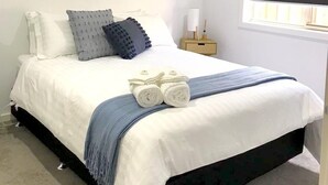 Un dormitorio con cama, almohadas, una mesita de noche con lámpara, un ventilador de techo y una ventana con persianas.