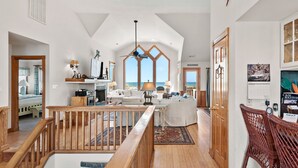 Ein geräumiger Wohnbereich mit hoher Decke, Holzboden, einer zentralen Sitzecke mit Sofa und Sesseln, einem Esstisch und großen Fenstern mit Blick auf den Strand.