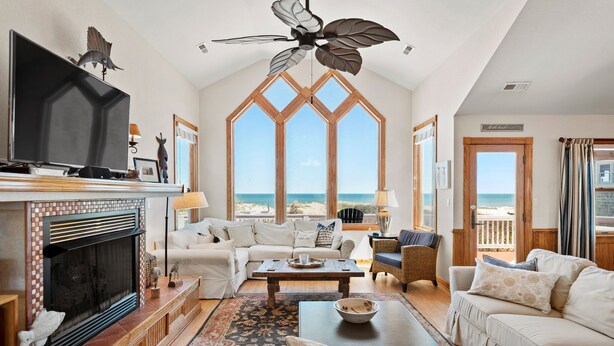 Ein geräumiges Wohnzimmer mit Kamin, einem großen Fernseher und Blick auf den Strand durch hohe Fenster.