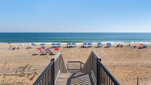 Ein Holzsteg führt zu einem Strand mit blau und rot gestreiften Sonnenschirmen.