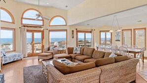 Un salon spacieux avec un grand canapé d’angle, une table à manger avec des chaises et un balcon offrant une vue sur la mer.