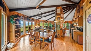 A wooden cabin interior with a dining area, kitchen, and a large window overlooking greenery.