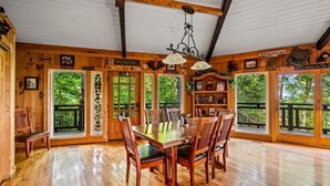 A wooden dining room with a large table and chairs, surrounded by windows with a view of trees.