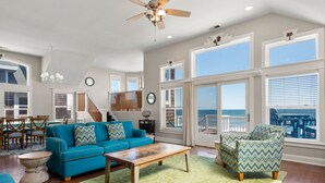 A spacious living room with a blue sofa, a wooden coffee table, and a green patterned rug. There is a staircase leading to a balcony with a view of the ocean. - Beach Breeze: Oceanfront, Pool, Hot Tub, Elevator (Nags Head)