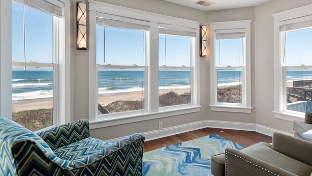 A room with large windows overlooking a beach, a patterned rug, and a sofa with a zigzag cushion. - Beach Breeze: Oceanfront, Pool, Hot Tub, Elevator (Nags Head)