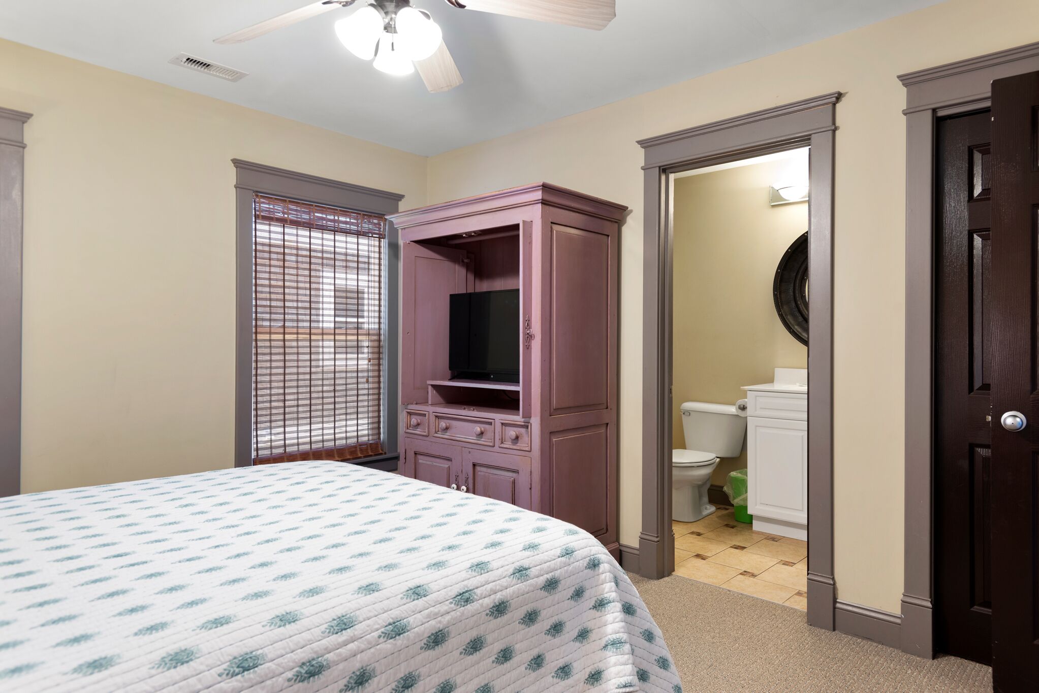 A bedroom with a bed, a wardrobe, a television, a window with blinds, and a bathroom visible through an open door.
