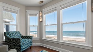A room with a large bay window overlooking the beach, a patterned armchair, and a wooden floor. - Beach Breeze: Oceanfront, Pool, Hot Tub, Elevator (Nags Head)