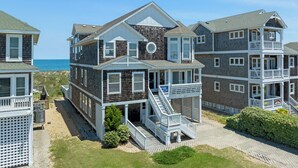 A multi-story beachfront house with a balcony, a staircase leading to the upper floor, and a view of the ocean. - Beach Breeze: Oceanfront, Pool, Hot Tub, Elevator (Nags Head)