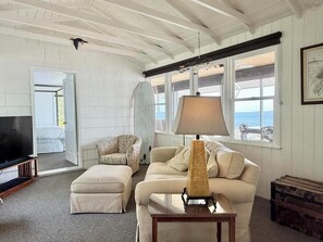Una sala de estar con una televisión, un sofá, una lámpara y vista al mar a través de amplios ventanales.