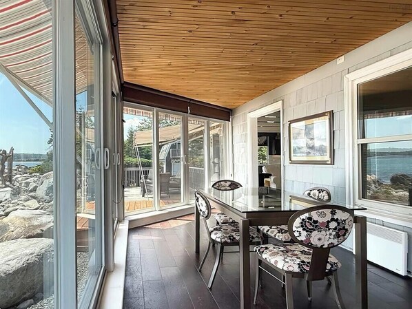 A modern dining area with a glass table and chairs, wooden ceiling, and large windows offering a view of the outdoors. - OCEAN SURF COTTAGE AT SUMMERVILLE BEACH (Summerville Centre)