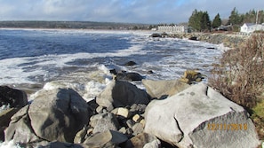 Una costa rocosa con olas rompiendo contra las rocas. Se ven casas al fondo, cerca del agua.