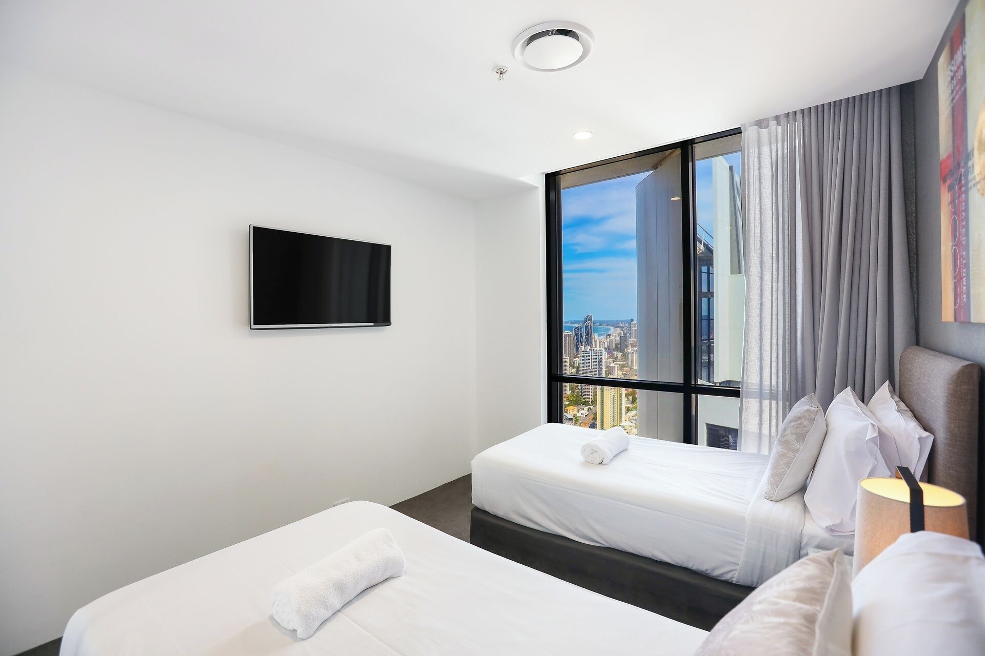 A modern hotel room with two beds, a flat-screen TV, and a large window offering a city view.