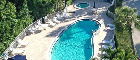 A large outdoor swimming pool with a curved design, surrounded by lounge chairs and umbrellas, set against a backdrop of lush greenery and a building.