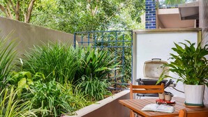 A patio with a wooden table and chairs, a grill, and lush greenery. - Stylish River Boulevard Apartment in Richmond (Richmond)
